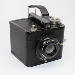 Könnte beinhalten: Eine schwarze Kodak Brownie Six-20 Boxkamera mit strukturierter Oberfläche, einem Tragegriff aus Leder und einem zentralen Objektiv. Das Objektivgehäuse ist mit den weißen Aufschriften "SIX-20", "BROWNIE" und "5 TO 10 FEET" versehen.