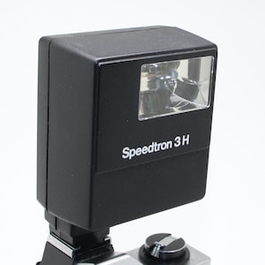 Op de afbeelding: Een zwarte Speedtron 3H flitser, gemonteerd op een camera. De flitser heeft een rechthoekige vorm met een helder voorpaneel. De tekst 'Speedtron 3H' is in het wit op de voorkant van de flitser gedrukt.