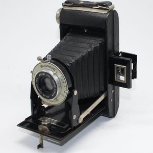 May include: A vintage black Kodak Brownie camera with a collapsible bellows design. The camera features a lens with aperture settings and a viewfinder. The camera is made of black leather and metal, with the word "Brownie" visible on the lens.