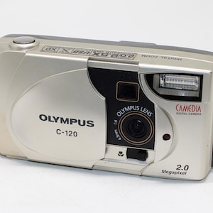 May include: A silver Olympus C-120 digital camera featuring a 2.0 Megapixel lens, a built-in flash, and a sliding lens cover. The front of the device displays the Olympus brand name, Camedia digital camera text, and a small flower icon.