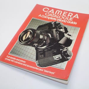 Puede incluir: Un catálogo de cámaras vintage con una portada roja que presenta una ilustración de una cámara Canon negra. El texto de la portada dice "CAMERA CATALOGUE A Complete World Guide". El texto adicional incluye detalles y precios de más de 140 cámaras y accesorios.