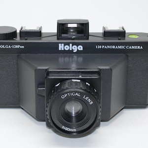 May include: Black Holga 120 Panoramic Camera with an optical lens and f=90mm. The camera has the text "Holga 120 Panoramic Camera" printed on the top. The camera has a viewfinder and a lens.
