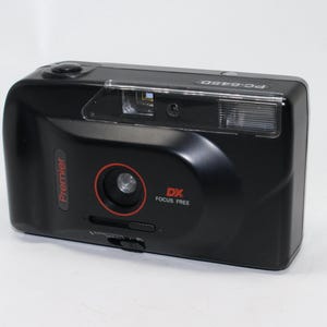 Może przedstawiać: Czarny aparat Premier PC-645D Focus Free. Aparat ma czerwone kółko wokół obiektywu i napis "DX FOCUS FREE" w kolorze czerwonym. Aparat ma lampę błyskową i mały przycisk na górze.