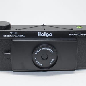 May include: Black Holga wide pinhole camera. The camera has the text "WIDE PINHOLE CAMERA" and "Holga" printed on the body. The lens has the text "WIDE PINHOLE" and "f/135" printed on it. The camera has a built-in level.