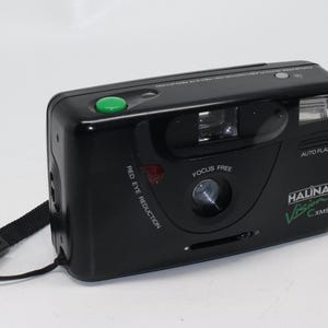 May include: A black Halina Vision CXMS 35mm film camera with a green button, red eye reduction, auto flash, and focus free lens. The camera has a black strap.