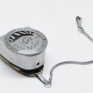 May include: A vintage light meter with a silver body and a circular dial featuring numbers and markings. It has a chain attached and is designed for measuring light levels in photography. The meter's dial includes settings for seconds and DIN.