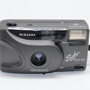 Puede incluir: Una cámara gris Miranda Solo Twin MD con flash incorporado y lente. La cámara tiene el texto "MOTORDRIVE" y "MIRANDA" impreso. Tiene una correa de muñeca.