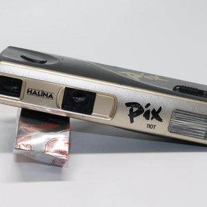 May include: A silver and black Halina Pix 110T film camera with a built-in flash. The camera is resting on a silver and orange film package.