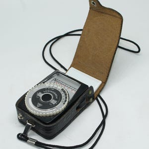 Könnte beinhalten: Vintage schwarzer Leningrad 4 Belichtungsmesser in einer Ledertasche mit schwarzem Riemen. Der Belichtungsmesser hat ein silbernes Zifferblatt mit schwarzen Zahlen und eine weiße Nadel. Die Ledertasche ist innen braun und hat einen Schnappverschluss.