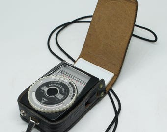 May include: A vintage black Leningrad 4 light meter in a leather case with a black strap. The light meter has a silver dial with black numbers and a white needle. The leather case is brown on the inside and has a snap closure.