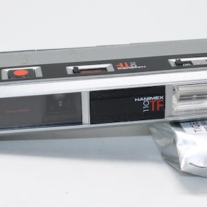 May include: A silver and black Hanimex 110 TF camera with a red shutter button and a built-in flash. The camera is paired with a silver film cartridge labeled "TIGER 200 ISO".