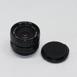 Puede incluir: Un objetivo de cámara negro Pentax-110 de 18 mm con una tapa de objetivo a juego. El objetivo tiene el texto "PENTAX-110 1:2.8 18mm" impreso. La tapa del objetivo tiene el texto "ASAHI PENTAX" impreso.