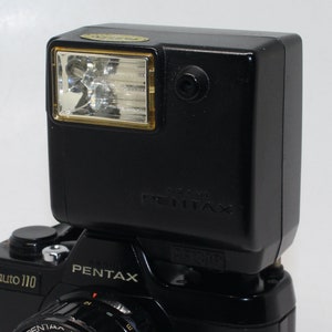 以下が含まれることがあります： 黒いペンタックスカメラのフラッシュユニットが、黒いペンタックスカメラに取り付けられています。フラッシュは、透明なレンズと金色のフレームが付いた長方形のヘッドを備えています。カメラ本体には「Pentax」と「auto 110」の文字があります。