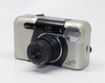 Pode incluir: Uma câmera Konica Z-up 70 Super de zoom de 35 mm a 70 mm de cor prateada com uma lente preta.