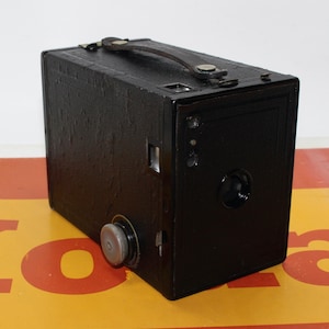 May include: A black vintage box camera with a leather carrying strap. The camera has a round lens and a small viewfinder on the top.