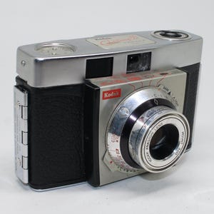 May include: Vintage Kodak camera with a silver top and lens, black body, and a red Kodak logo. The camera has a silver lens ring with black and white markings. The camera is a classic film camera.
