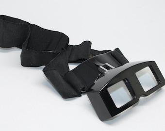 May include: Black 3D glasses with a black strap. The glasses have a rectangular shape with two square lenses. The strap is made of a thick, woven material and is attached to the glasses with a buckle.