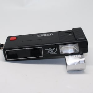 May include: Black Cobra 20 Pocket Camera with a red button and a silver flash attachment. The camera has a rectangular shape with a lens and a flash. The camera is on a white surface.