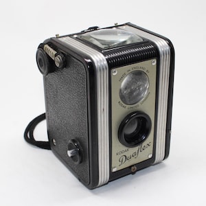 Könnte beinhalten: Eine Vintage Kodak Duaflex Boxkamera mit schwarzem, texturiertem Gehäuse und silbernen Metallakzenten. Die Vorderseite zeigt eine silberne Platte mit dem Text Made in England by Kodak Limited London und dem Wort Kodak Duaflex in Schreibschrift.