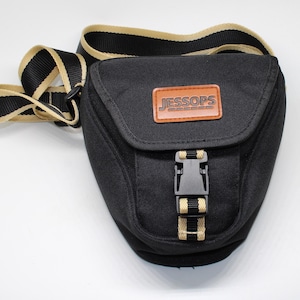 Può includere: Una borsa per fotocamera nera con tracolla regolabile beige e nera. La patta anteriore presenta una toppa rettangolare marrone con il testo JESSOPS impresso in nero. Una chiusura con fibbia in plastica nera fissa la patta anteriore.