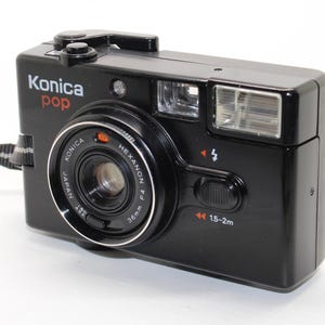 May include: A black Konica Pop 35mm film camera featuring a Hexanon 36mm f/4 lens, a built-in flash unit, a viewfinder, and a shutter button. The front of the camera displays the text Konica Pop, Japan, and distance markings of 1.5-2m.