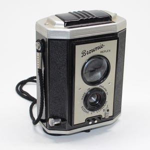 Könnte beinhalten: Eine schwarz-silberne Kodak Brownie Reflex Kamera mit strukturiertem Gehäuse, einem Sucher an der Oberseite und einem schwarzen Trageriemen. Die Frontplatte trägt die Aufschrift Brownie REFLEX und Made in England by Kodak Ltd London.