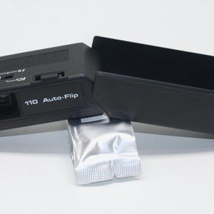 May include: Black Halina 110 Auto-Flip camera with a red button and a black case. The camera has the word "HALINA" printed on the front. A silver film pack is also included.