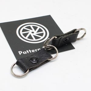 May include: Two black textured leather key fob connectors featuring silver-toned metal split rings on each end and a central black snap button closure. The items are positioned over a black square card with a white circular wheel graphic and the text Pattern.