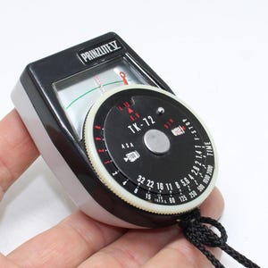 May include: A Prinzlite V light meter featuring a black and white casing with a circular dial labeled TK-72. The device includes a needle display, ASA settings, F-stop markings, and time indicators, with a black cord attached to the base.