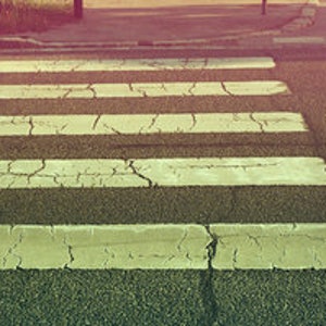 May include: A white pedestrian crossing painted on a grey asphalt road surface. The crossing is cracked and worn.