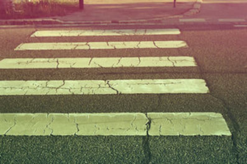 May include: A white pedestrian crossing painted on a grey asphalt road surface. The crossing is cracked and worn.