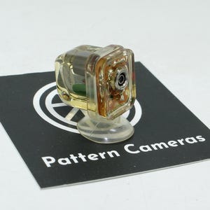 Peut inclure: Un petit appareil photo transparent avec un corps transparent et un objectif. L'appareil photo est monté sur une base transparente et arrondie. Il repose sur une carte noire avec le texte blanc "Pattern Cameras".