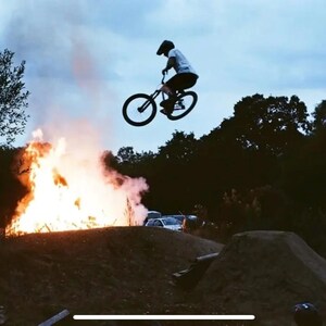 Puede incluir: Una persona realiza un truco en una bicicleta de monta&ntilde;a negra frente a una hoguera con humo elev&aacute;ndose en el aire. La persona lleva un casco y una camisa blanca. El fondo incluye &aacute;rboles y un camino de tierra.