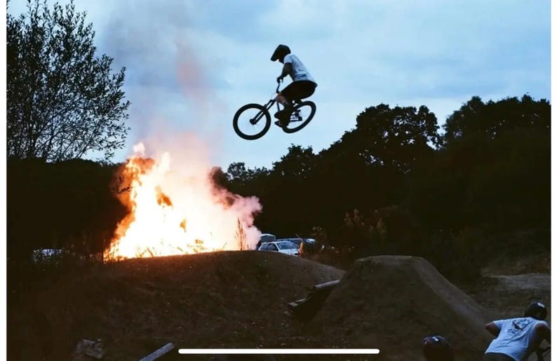 Puede incluir: Una persona realiza un truco en una bicicleta de monta&ntilde;a negra frente a una hoguera con humo elev&aacute;ndose en el aire. La persona lleva un casco y una camisa blanca. El fondo incluye &aacute;rboles y un camino de tierra.