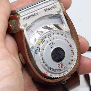 May include: A vintage Hanimex Sekonic light meter encased in a brown leather protective sleeve. The device features a circular silver dial with black numerical markings for ASA, light value, and aperture settings, plus a clear plastic top cover.