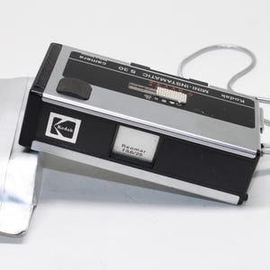 以下が含まれることがあります： 黒と銀色のKodak Mini-Instamatic S 30カメラ。Reomar f/5.6 25mmレンズ、黒いシャッターボタン、白いスライド式フォーカススイッチ、銀色の金属製ネックチェーンを備え、長方形の銀色のアルミホイルパケットが付属しています。