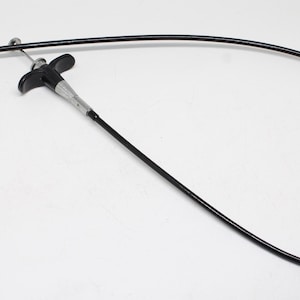 May include: A black camera shutter release cable with a silver metal tip at one end and a black plastic plunger mechanism with a silver collar at the other end. The flexible cable is curved into a large loop on a plain white background.