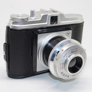 May include: A vintage Agfa Isola camera featuring a black textured body with silver metal accents. The lens barrel displays distance markings in both feet and metres, with red and black text. The top plate includes a silver winding knob.