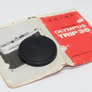 Op de afbeelding: Een zwarte Olympus lensdop ligt bovenop de handleiding voor een Olympus Trip 35 camera. De handleiding is open en toont een diagram van de camera. De omslag is rood en wit, met de woorden "Olympus Trip 35" erop gedrukt.