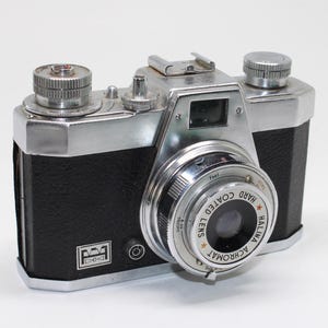 May include: A vintage 35mm film camera with a black textured body and silver metal top plate. The lens barrel is marked with the text Halina Hard Coated Lens Achromat. It features a top-mounted viewfinder, film advance knob, and shutter button.
