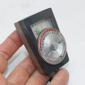 May include: A vintage Sekonic light meter with a gray body and a silver dial. The dial features numbers and markings in red and white, with a needle indicator. The meter has the text "EKONI" and "SEKONIC" on it.