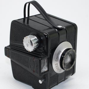 Puede incluir: Una cámara Ilford Envoy negra vintage con un anillo de lente plateado y un asa de cuero negro. La cámara tiene un cuerpo texturizado y un dial cromado. El anillo de la lente dice "Ilford Envoy Optimax Lens".