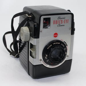 May include: A black and silver Kodak Brownie Bull's-Eye camera with a black strap. The camera has a red "Kodak" logo and a "Kodak Twindar Lens" with a long and instant setting.