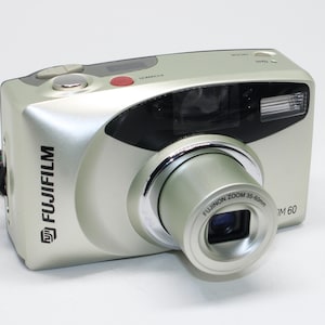 May include: Silver Fujifilm M60 digital camera with a zoom lens. The camera has a built-in flash and several buttons on the top. The lens is labeled "FUJINON ZOOM 35-60mm". The camera is on a white background.