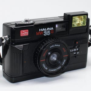May include: Black Halina MW35G camera with a Halinar 38mm lens. The camera has a built-in flash and a red and black strap. The camera has the word "MOTOR" and the Halina logo on the front.