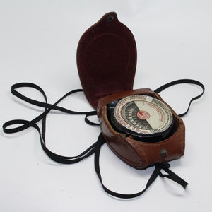 Può includere: Una custodia in pelle marrone vintage con un cinturino nero e un esposimetro circolare con un quadrante bianco e segni neri. L'esposimetro ha un logo rosso e bianco che dice "LUMI" e "SPEED".