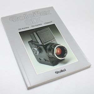 May include: A grey book titled "Rolleiflex SIX" featuring a photograph of a black camera with a prominent lens and a red interior. The book also has the words "Die Kamera - The Camera - L'Appareil" printed on it.