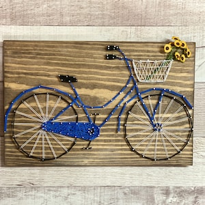 May include: Blue bicycle string art with white string spokes and a white wire basket with yellow flowers. The bicycle is mounted on a brown wooden background.