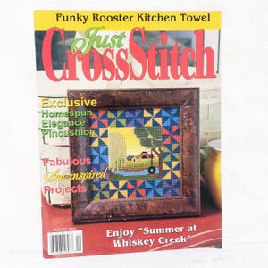 May include: A magazine cover with the title "Just CrossStitch" in red and green. The cover features a framed cross-stitch design with a landscape scene. Text includes "Funky Rooster Kitchen Towel" and "Enjoy 'Summer at Whiskey Creek.'"