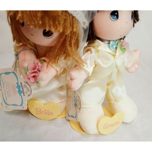 May include: A pair of plush wedding dolls, a bride and groom, with the words "Bride" and "Groom" on their bases. The bride is holding a pink flower bouquet. The dolls are part of the "Little Miss & Mister" collection.